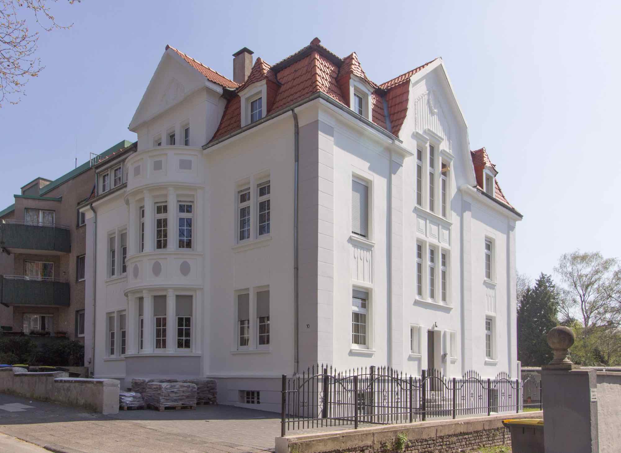 Helles Mehrfamilienhaus mit rotem Ziegeldach und Erkern vor blauem Himmel
